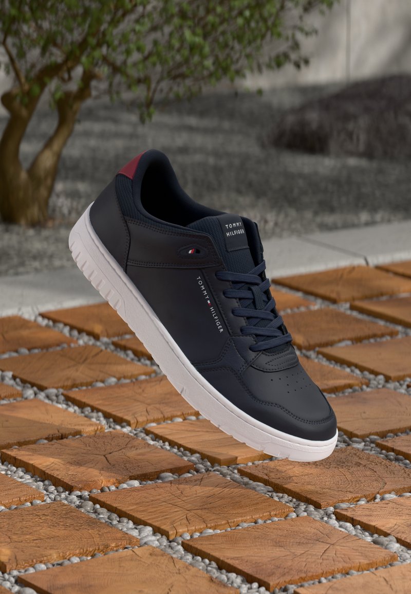 Baskets Tommy Hilfiger noires et rouges avec une semelle blanche reposant sur des dalles carrées en bois et de petits cailloux en extérieur.