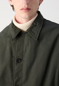 Olivengrøn, letvægtsfrakke med krave og sorte knapper; båret over en cremefarvet, ribbebetrukket turtleneck sweater. Glat, mat tekstur.