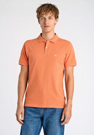 Oranssi puuvillahaalari, jossa on klassinen kaulus, kaksi nappia ja pieni logo rinnassa. Yhdistetty sinisiin farkkuihin.