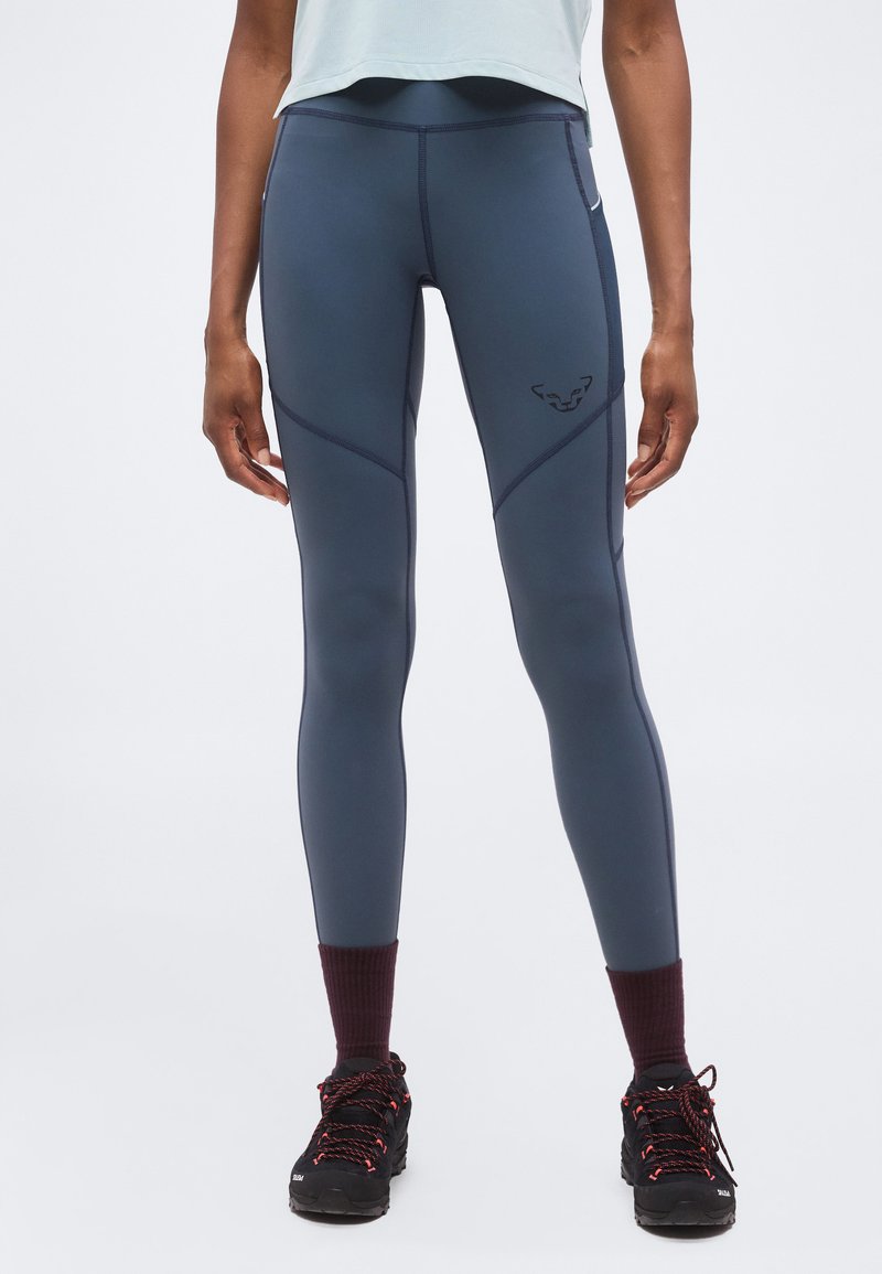 Άτομο που φοράει στενά μπλε-γκρι κολάν με μαύρο λογότυπο στον μηρό, συνδυασμένο με σκούρα ορειβατικά παπούτσια και μπορντό κάλτσες, στέκεται σε απλό φόντο.