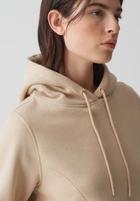 Felpa con cappuccio beige realizzata in un tessuto morbido e testurizzato, con cordino e dettagli delle cuciture sottili. Inquadratura ravvicinata, con focus sul colletto.