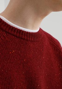 Punainen villapaita, jossa on tyylikäs neulekuvio. Ominaisuuksina pyöreä ribbineck, kuvattuna olkapäästä kaula-alueeseen.