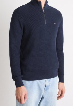 Pullover blu navy con una superficie texturizzata, colletto alto e zip frontale. Polsini e orlo a coste, con un piccolo logo sul petto.