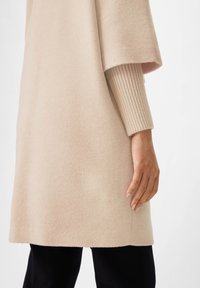 Beigen villaisen tunika, jossa on pehmeä koostumus, väljä istuvuus ja resorireunaiset hihat, yhdistettynä mustiin housuihin.