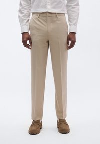 Pantalones beige entallados con una textura suave, con corte recto y bolsillos laterales, combinados con zapatos marrones sin cordones.