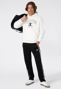 Felpa bianca con logo "Champion" e accenti neri, abbinata a pantaloni sportivi neri con strisce laterali bianche e logo. L'individuo porta una borsa nera.