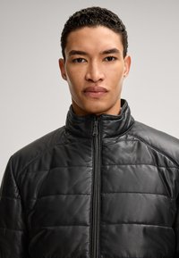 Schwarze Lederjacke mit gestepptem Design, hohem Kragen und Frontreißverschluss. Glatte Textur, taillierte Form und keine sichtbaren Verzierungen.