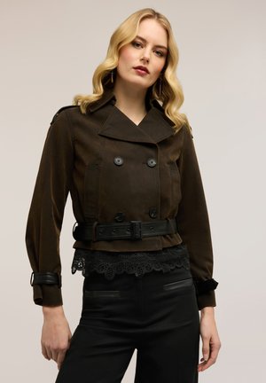 Junge Frau mit blonden Haaren trägt eine dunkelbraune Jacke mit Gürtel über einem schwarzen Spitzenoberteil und schwarzen Hosen, stehend vor einem einfarbigen Hintergrund.