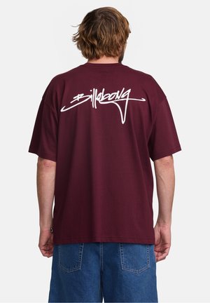 Mężczyzna z falującymi brązowymi włosami, ubrany w luźny bordowy T-shirt z białym napisem "Billabong" na plecach i niebieskie szorty dżinsowe, widziany od tyłu.