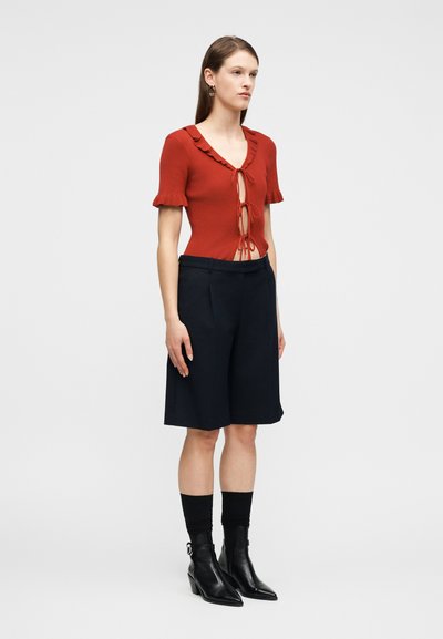 Γυναίκα που φοράει κορυφή με κοντά μανίκια σε σκουροκόκκινο χρώμα με δεσίματα μπροστά, μαύρα σορτς μέχρι το γόνατο, μαύρες κάλτσες και μαύρα μποτάκια αστραγάλου, στέκεται μπροστά σε απλό φόντο.