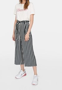 T-shirt blanc avec texte rose, culottes rayées noir et blanc, et baskets blanches avec accents roses. Tenue décontractée avec une taille nouée.