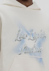 Sweat à capuche couleur crème avec le texte "Low Lights Studios" en argenté brillant sur un fond dégradé bleu, et une poche kangourou à l'avant.