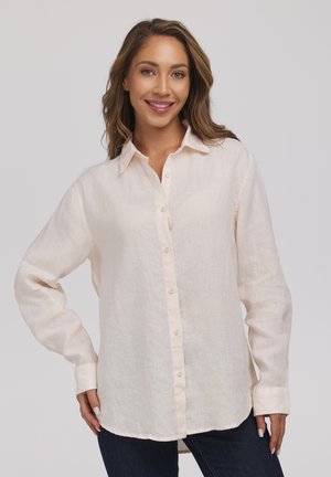 Kobieta z długimi falującymi włosami, ubrana w luźną, kremową koszulę z guzikami i ciemne jeansy, uśmiechająca się z jedną ręką na biodrze.