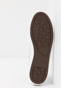 Białe płócienne sneakersy z brązową gumową podeszwą z diamentowym wzorem i czarnym logo. Z boku znajdują się oczka na sznurowadła.