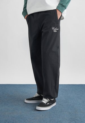 Person iført sorte Carhartt WIP bukser med lommer og sorte canvas sneakers med hvide snørrebånd, stående på blå tæppebelagt gulv.