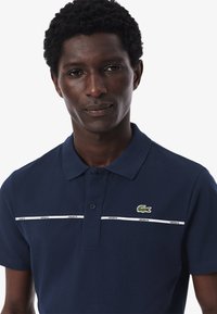 Mörkblå pikétröja av texturerad tyg med en grön krokodil-logotyp och två vita horisontella ränder med "LACOSTE" tryckt på dem.