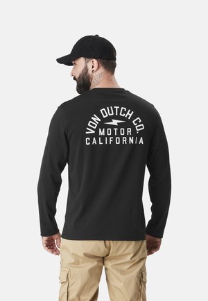T-shirt noir à manches longues arborant un texte graphique blanc au dos indiquant "VON DUTCH CO. MOTOR CALIFORNIA", associé à un pantalon cargo beige.