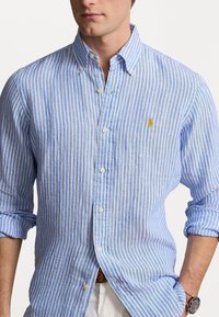 Světle modrá a bílá pruhovaná košile na knoflíky vyrobená z látky s texturovaným povrchem; má malé žluté logo a klasický design límce.