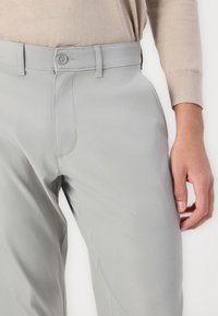 Ljusgrå byxor med en slät yta, utrustade med knappstängning och hällor för bälte. Närbild som framhäver framsidan och en hand.