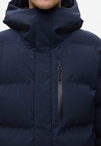 Námořnická puffer bunda s vysokým límcem a kapsou na zip na hrudi. Hladká textura, prošívaný design a nastavitelná kapuce.