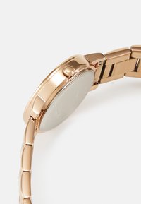 Auksinės spalvos metalinis laikrodis su apvaliu korpusu, lygia danga ir tekstūruota karūnėle, turintis poliruotą apyrankę su skirtingų dydžių grandinėmis.