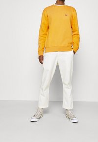 Oranje sweatshirt met ronde hals, geribbelde boorden, gecombineerd met witte broek en lichtgrijze hoge sneakers. Eenvoudig ontwerp, minimale branding.