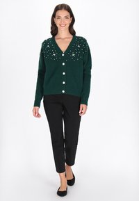 Zielony sweter zapinany na guziki z dekoltem w serek, ozdobiony koralikami na ramionach, w komplecie z czarnymi spodniami.