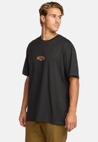 Camiseta de algodón negra con cuello redondo y mangas cortas. Presenta un logotipo central en naranja y amarillo con un diseño curvado.