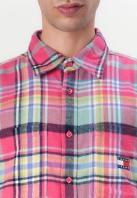 Pink and green plaid flannel shirt with button-up front, rounded collar, chest pocket, and logo patch. Features small pink buttons.