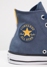 Nízko na kotníku modré tenisky s texturovaným povrchem, s žlutou a modrou nášivkou se hvězdou, bílou podrážkou a šedým akcentem.