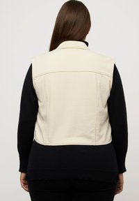 Gilet crema realizzato in tessuto morbido con colletto alto, caratterizzato da dettagli a coste neri nella parte inferiore e maniche lunghe sotto.
