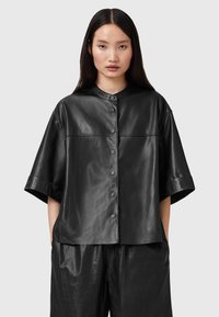 Black leather boxy shirt with a round collar, short sleeves, and button front. Smooth texture with visible stitching and minimal design.