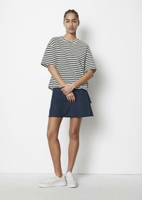 Kvinne står mot en hvit vegg iført en svart og hvit stripete skjorte, marineblåt skjørt, hvite joggesko, med håret i fletter.