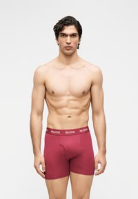 Άνδρας χωρίς πουκάμισο με κοντά σγουρά σκούρα μαλλιά, ντυμένος με κόκκινα εσώρουχα Hollister, στέκεται σε απλό φωτεινό φόντο.