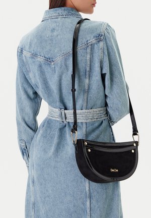 Mujer con chaqueta vaquera azul claro con cinturón, llevando un bolso de hombro negro DeeZee con herrajes dorados y correa ajustable.