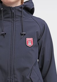 Marineblauwe zip-up jas met capuchon. Bevat een rood en wit geborduurd logo op de borst. Verstelbare koordjes en zijzakken.