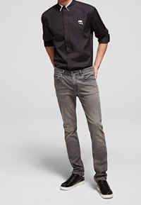 Černá košile na knoflíky s bílým límečkem, šedé džíny a černé tenisky. Výrazný detail loga na košili. Slim-fit design. Hladká textura.