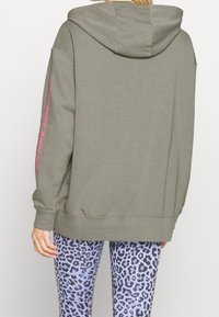 Coupe oversize en olive avec une capuche, présente un logo Nike rose sur la manche gauche. Associée à des leggings violets à motifs léopard.