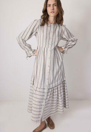 Žena s vlnitými hnedými vlasmi, oblečená v dlhých bielych šatách s vertikálnymi sivými pruhmi a hnedými tkávanými balerínkami, pózujúca s rukami v bok.