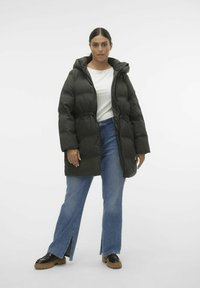 Manteau doudoune noir avec une capuche, taille cintrée et manches longues. Porté sur une chemise blanche et associé à un jean large bleu clair et des chaussures noires.