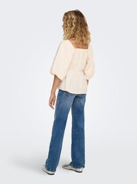 Kremīgs blūzes ar pūtīšu piedurknēm, kvadrātveida izgriezumu, teksturētu audumu un savāktu muguras detaļu, kombinēta ar augsta vidukļa izbalējušām džinsa biksēm.