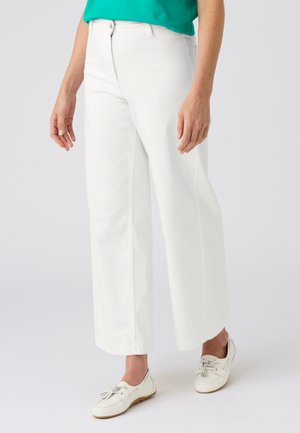 Mujer con pantalones blancos de pierna recta, mocasines blancos con borlas y una blusa verde, de pie contra un fondo claro y sencillo.