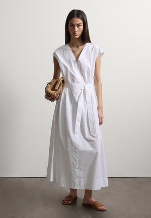 Femme portant une robe portefeuille blanche sans manches avec une ceinture, tenant une pochette tissée et portant des sandales marron à enfiler sur un fond uni.