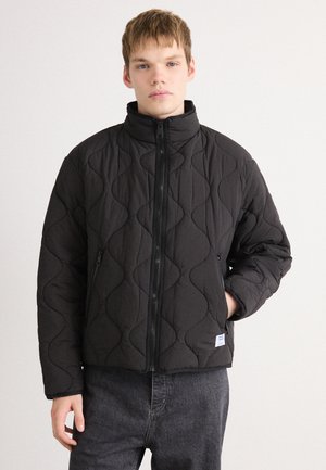 Jonge man in een zwarte gewatteerde zip-up jas met hoge kraag en zijzakken, staand tegen een effen lichte achtergrond.
