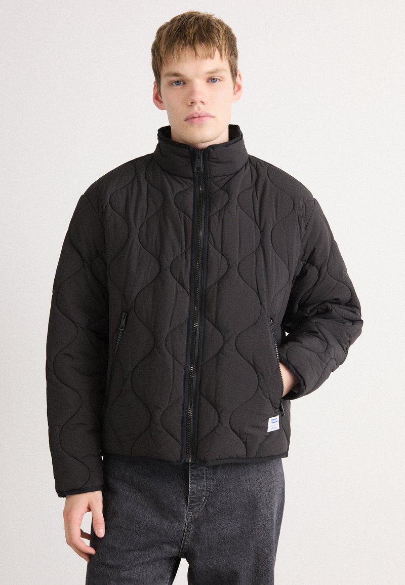 Young man wearing a black quilted zip-up jacket with high collar and side pockets, standing against a plain light background.