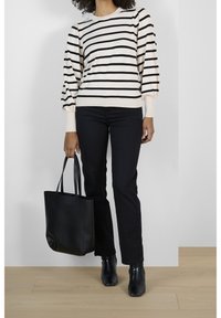 Nainen, joka on pukeutunut musta-valkoiseen raidoitettuun pitkähiharaiseen paitaan, mustiin housuihin, mustiin nilkkabootseihin ja kantaa mustaa nahkaista olkalaukkua.