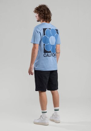 Camiseta de algodón azul claro con un gran gráfico de una flor azul y el texto "CALIFORNIA" en la parte de atrás, combinada con pantalones cortos negros y zapatillas blancas.