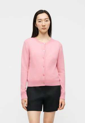 Femme aux longs cheveux foncés portant un cardigan rose boutonné et un short noir, debout devant un fond clair uni.