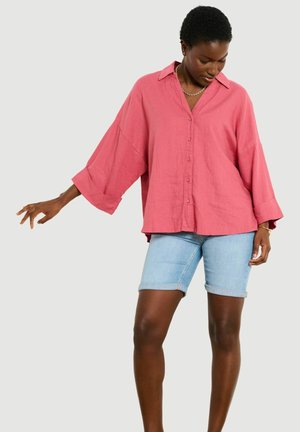 Žena oblečená v voľnej ružovej košeli na gombíky a svetlomodrých rifľových kraťasoch do kolien, stojaca s jednou natiahnutou rukou a pozerajúca sa nadol.