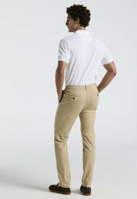 Chinos beiges légers associés à un polo blanc uni. Le pantalon dispose de poches arrière avec des boutons, et la tenue est complétée par des chaussures marron.